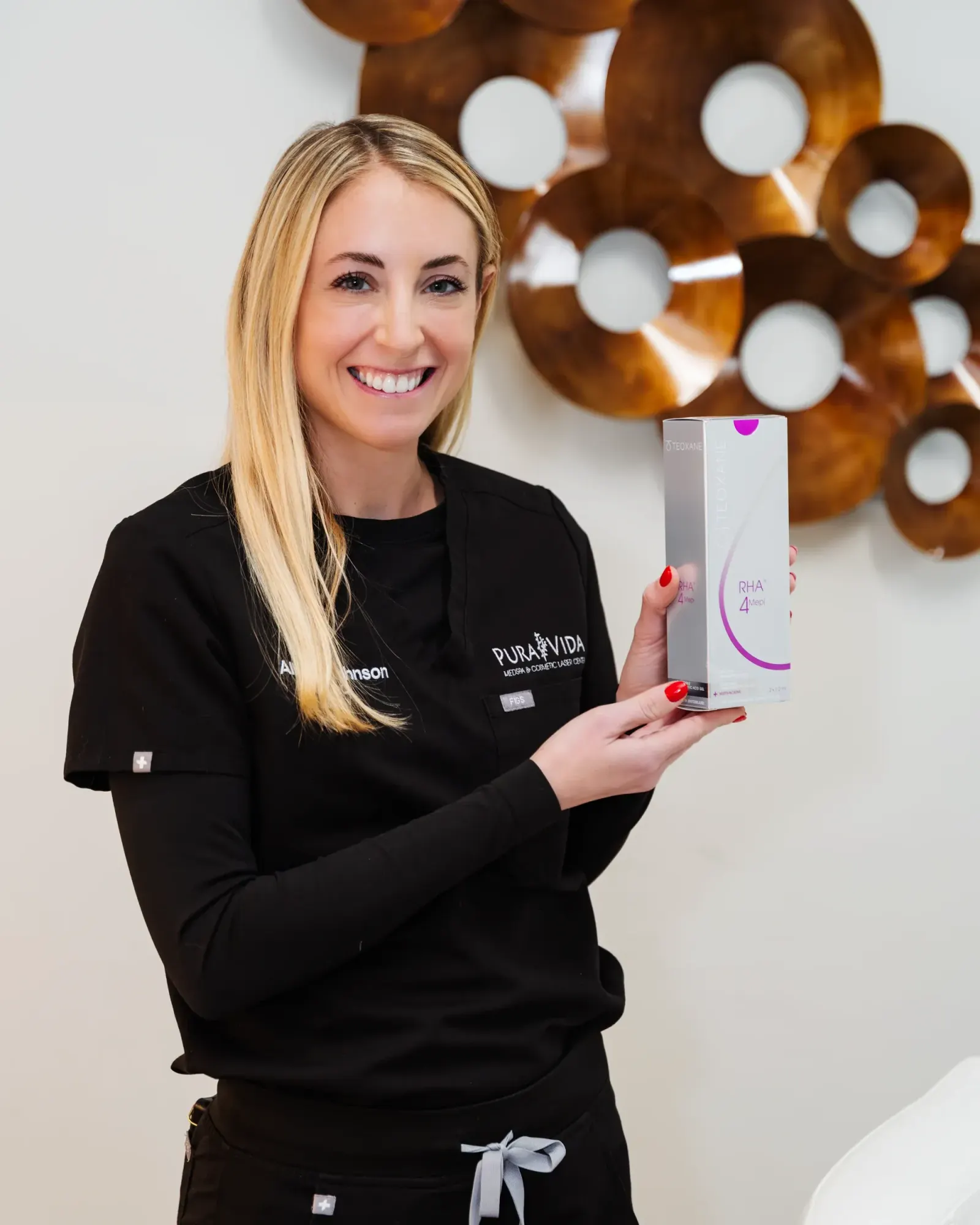 Smiling provider in black scrubs holding a dermal filler product box