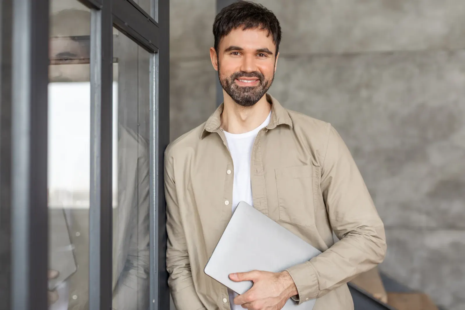 Smiling bearded man in beige shirt holding a laptop by a window