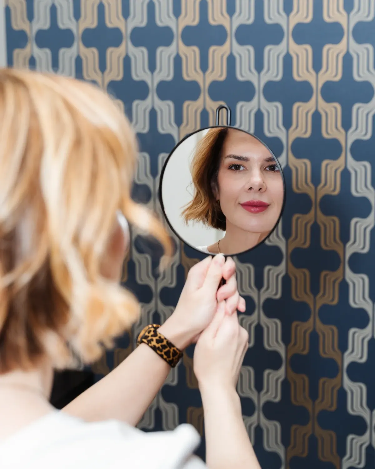 Woman checking her reflection in a handheld mirror against patterned wallpaper