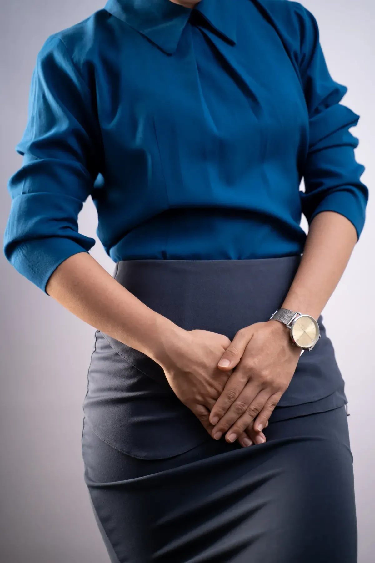 Woman in blue blouse holding hands over lower abdomen against neutral background