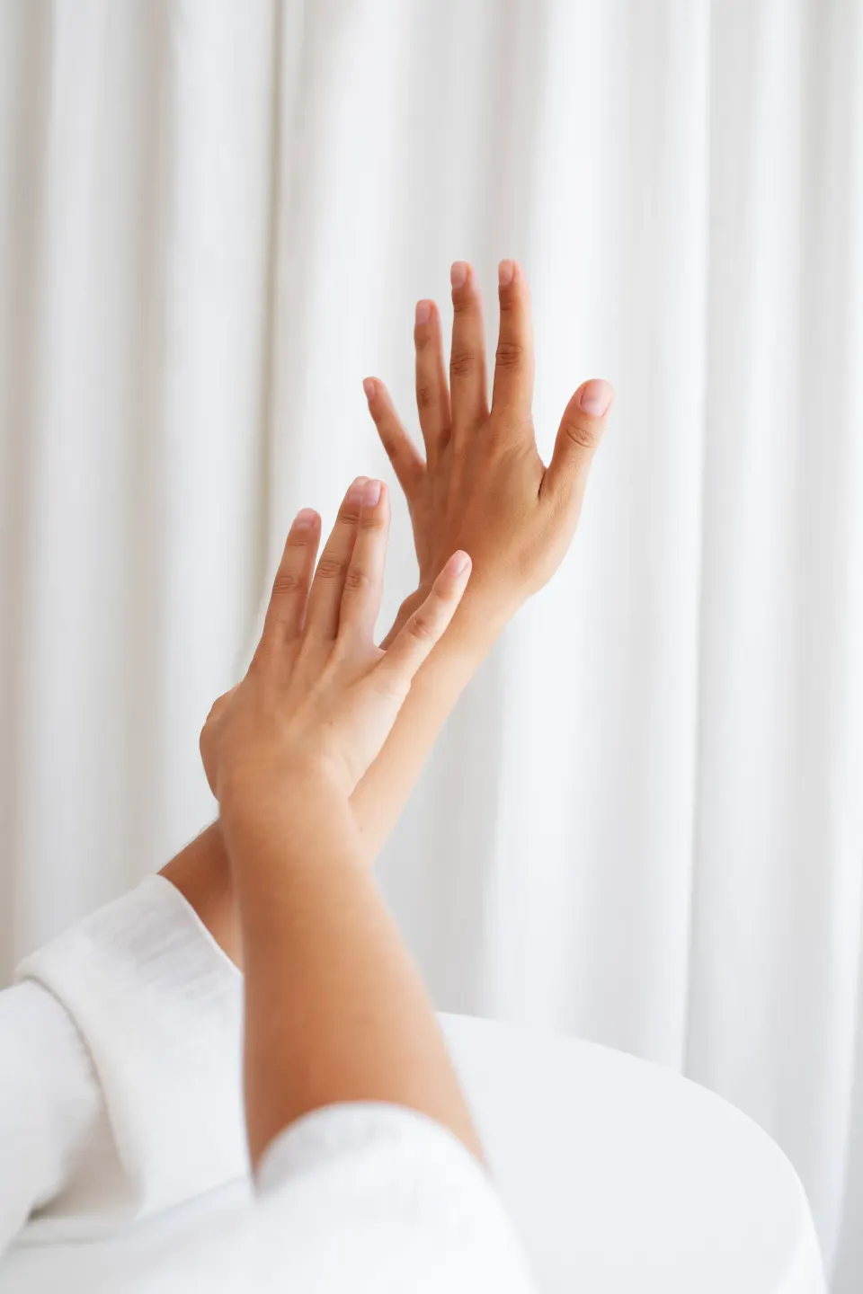 Woman's raised hands with smooth skin against a soft white curtain
