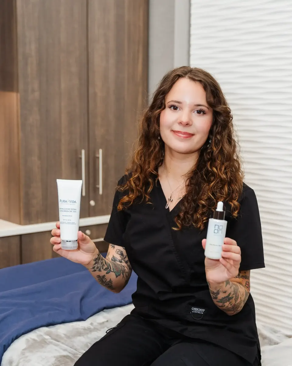 Smiling esthetician holding skincare cleanser and serum bottles in treatment room