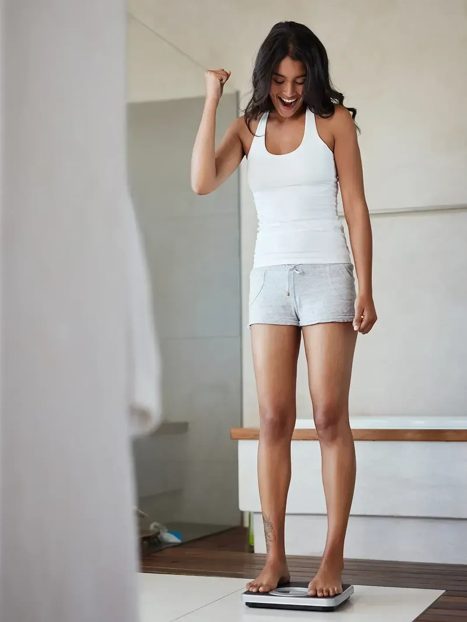 Woman in white tank top and shorts cheering while standing on a bathroom scale