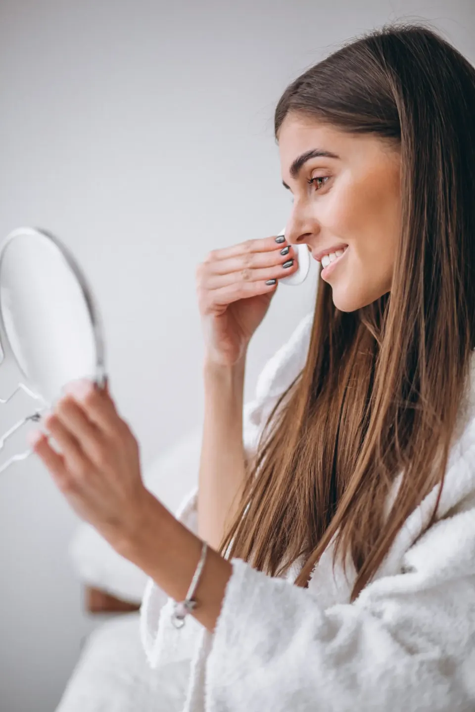 Woman in robe cleansing her face with a cotton pad before a mirror